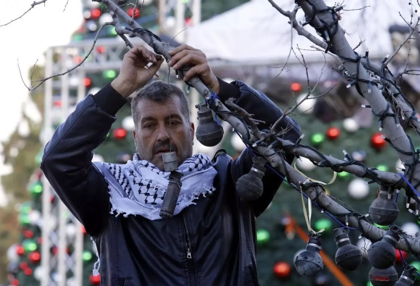 A person wearing a keffiyeh secures tear gas canisters to a tree, with normal Christmas ornaments visible on trees behind him