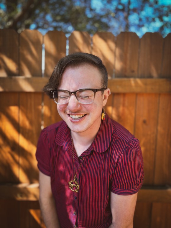 Photo of Avery, a white person with glasses and short brown hair, standing outside in a scarlet shirt. They are laughing with their eyes shut.