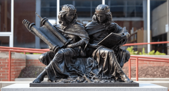An outdoor statue of two women sitting side by side. Both are robed and crowned. One holds a large torah scroll; the other an open Bible. They are smiling and looking at one another's texts.